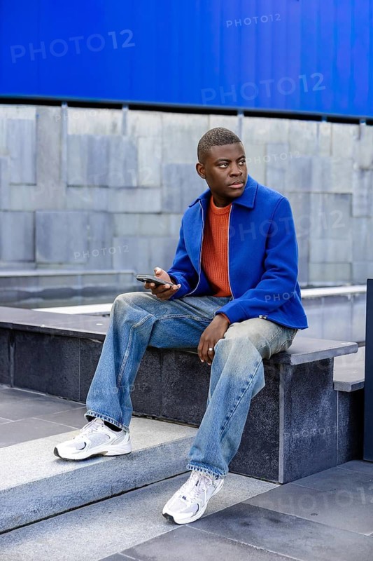 A young Black man sits on concrete steps, wearing a blue jacket and orange shirt. He holds a smartphone and looks thoughtfully into the distance