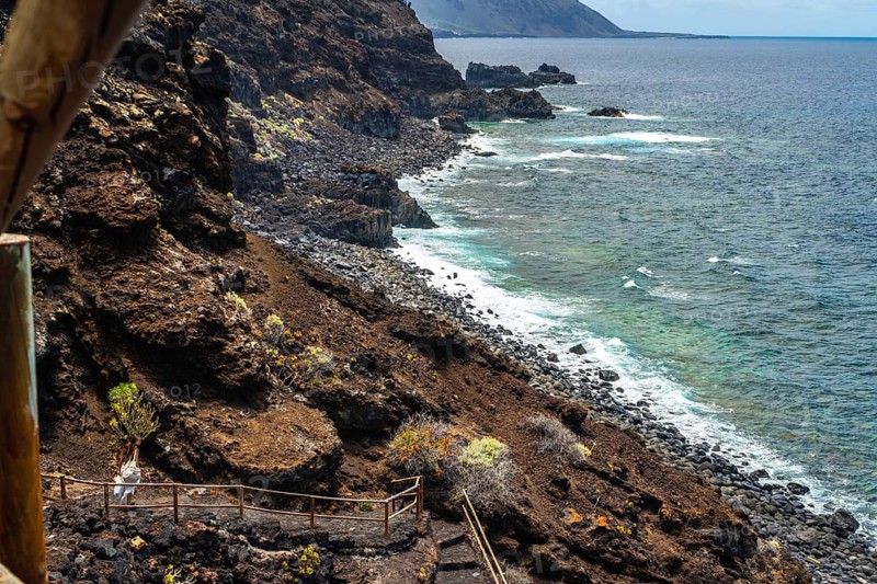 Couple walking path along rugged coastline with dark volcanic cliffs meeting the turquoise atlantic ocean on a sunny day in el hierro, canary islands