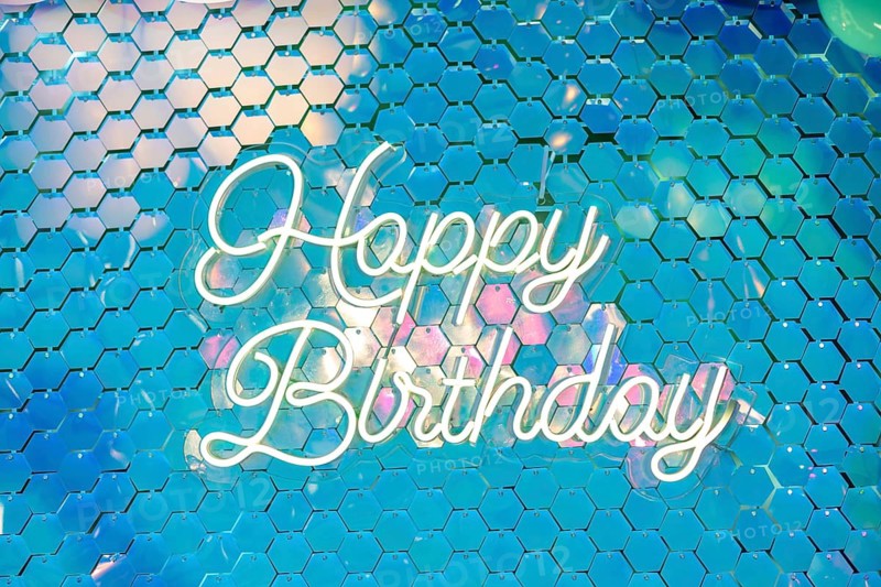 Colorful background with a neon sign that reads 'Happy Birthday'. The design features a shimmering, iridescent surface with hexagonal patterns