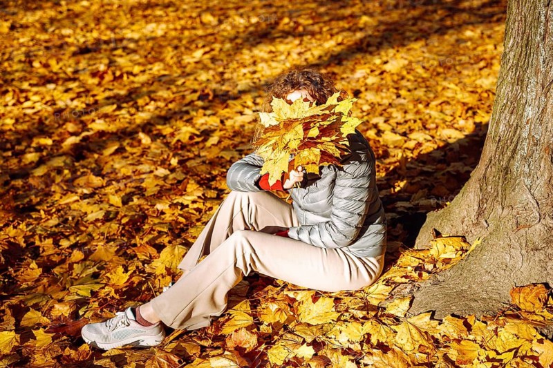 Woman holding autumn leaves in front of face in park. A person sits on the ground in a thick layer of autumn leaves, holding a bunch of yellow foliage that covers their face. White sneakers and a gray jacket stand out against the warm seasonal colors. The scene conveys fall atmosphere, playful concealment, cozy outdoor leisure, mindfulness, and the textured beauty of autumn nature