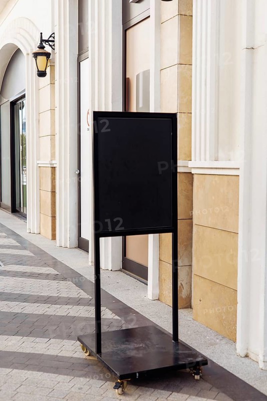 Mockup, black sidewalk sign stand is positioned on a bustling city street. Surrounding shops and palm trees create a lively atmosphere, suggesting an inviting environment for passersby