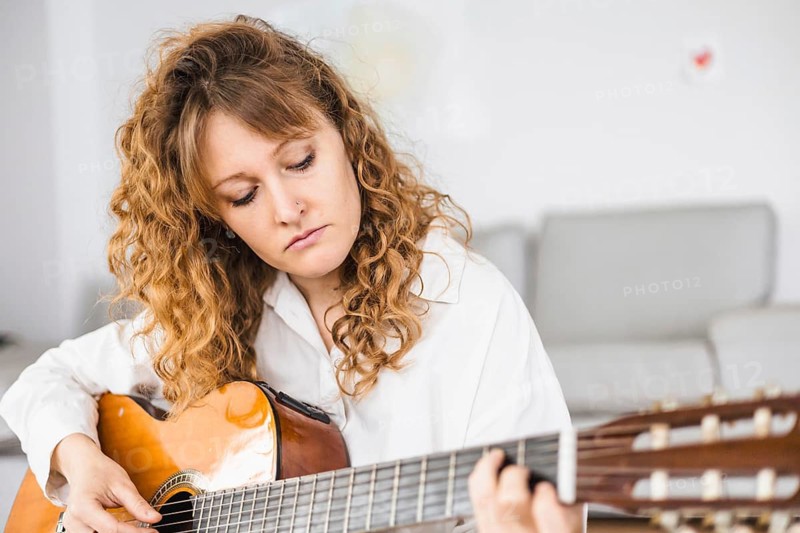 Curly-haired woman intently playing a guitar, wearing a white shirt
