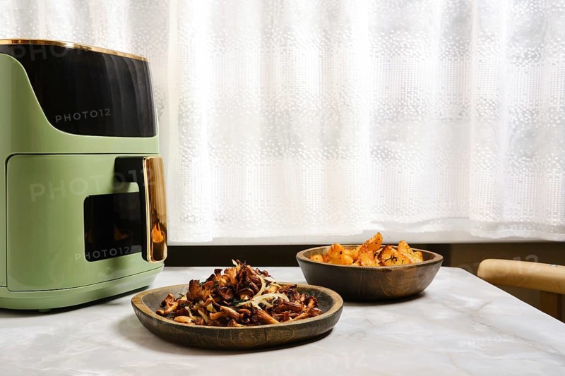 Air fryer sits on table with two wooden bowls nearby. One bowl contains roasted chanterelle mushrooms while the other has potato wedges