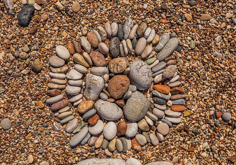 A circular arrangement of smooth pebbles on a sandy surface. The stones vary in size and color, creating a natural pattern. The scene is tranquil and earthy