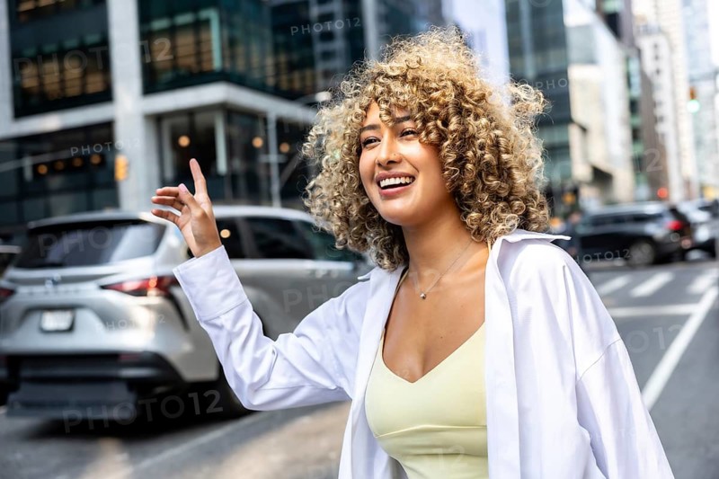 Young woman with curly hair smiling and raising her hand to hail a taxi, embodying urban lifestyle and happiness on a bustling manhattan street in new york city