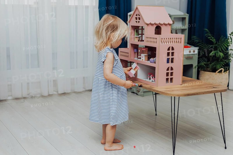 Child in a blue dress standing in a bright room playing with a dollhouse on a wooden table