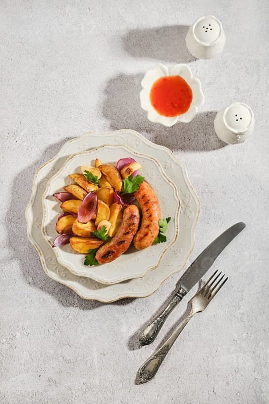 Top view of plate with grilled sausages and roasted potatoes. Green leaves decorate meal. Two small containers of sauce sit nearby. Fork and knife rest beside the plate