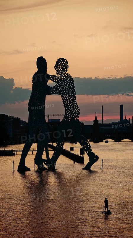Person on stand-up paddle in front of Molecule Man, Molecule Men, sculpture, figures in the water of the Spree against an impressive evening sky and urban environment, sunset, Berlin