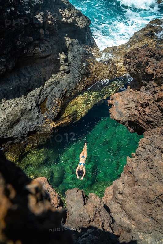 Woman floating peacefully on her back in a natural ocean pool with clear emerald water, surrounded by rugged volcanic rock formations on the coast of el hierro island