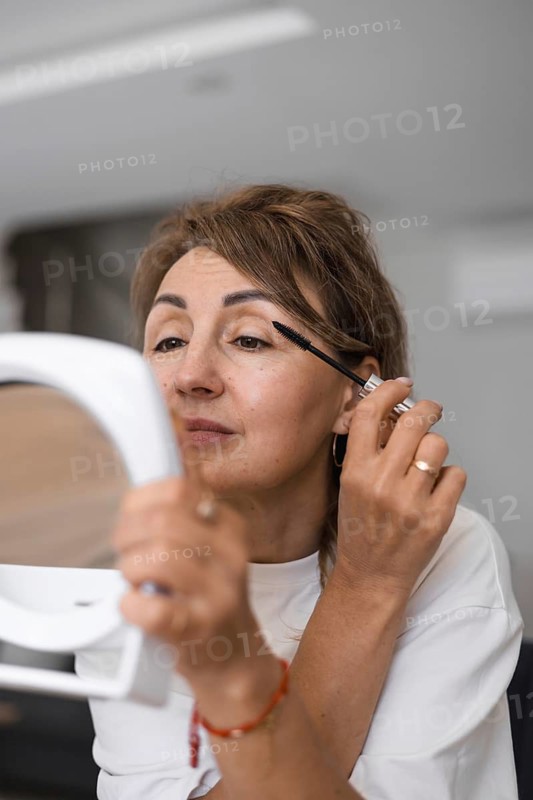 Portrait of mature woman sits at a wooden table, focused on applying mascara while looking into a mirror. Sunlight filters through the window, illuminating the space filled with greenery