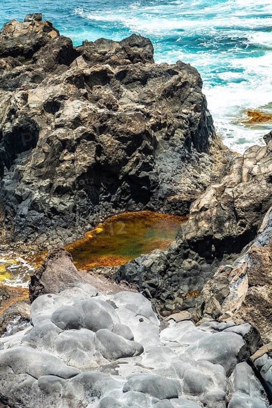 Jagged volcanic rock formations creating a natural pool with orange hued nutrient rich water, contrasting with the deep blue atlantic ocean waves crashing on the coast of el hierro island, spain