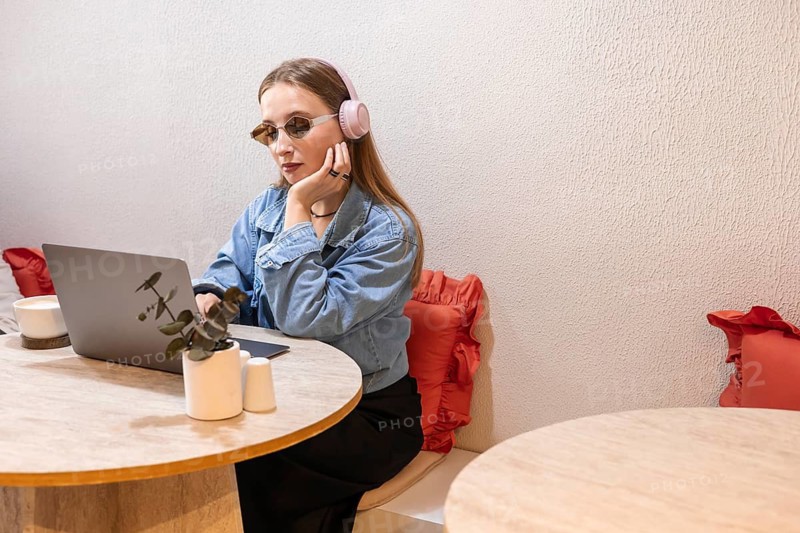 Young Caucasian woman with long blonde hair wearing pink headphones and sunglasses, working on a laptop in a modern indoor setting