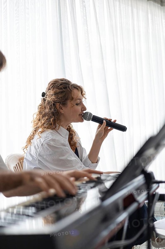 A young Caucasian woman with curly hair sings into a microphone. A hand plays a keyboard in the foreground. Bright, airy home setting with white curtains