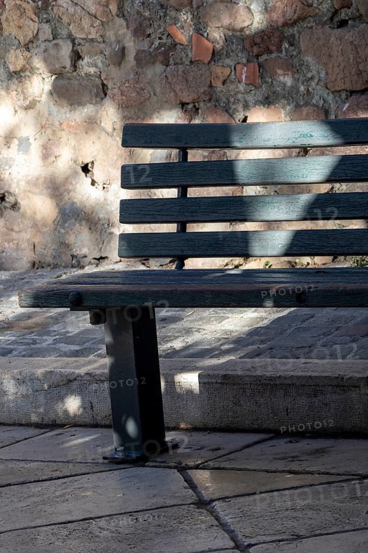 A dark green wooden bench stands beside a rough stone wall and steps in dappled light, Saint-Tropez, Var, Provence-Alpes-Cote d'Azur, France