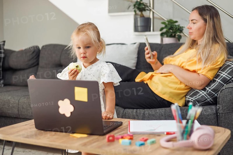 Young blond European girl eating sandwich with laptop on table. Pregnant blond European woman sitting on couch using smartphone. Home environment