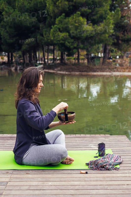 A young Caucasian woman with long brown hair sits cross-legged on a green yoga mat by a lake. She holds a singing bowl and appears to be meditating