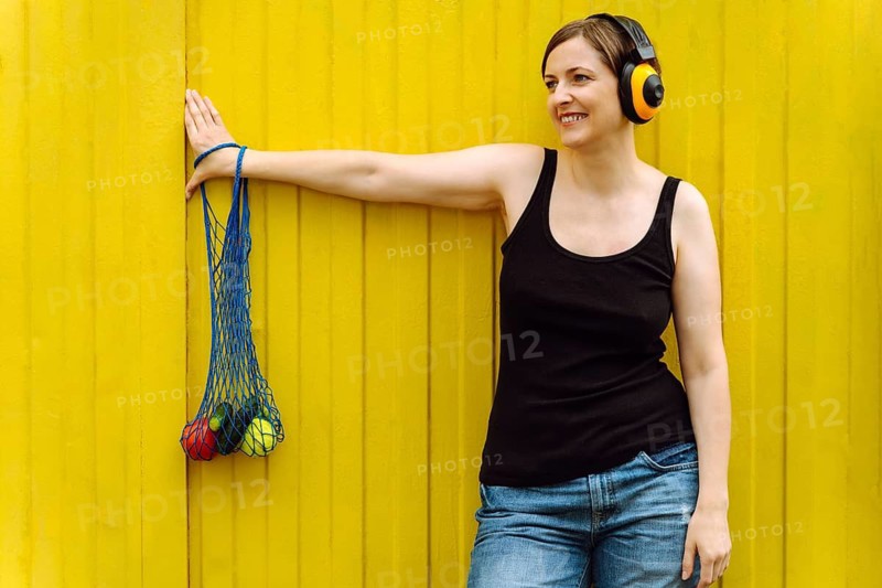 Smiling woman with yellow headphones carrying eco grocery bag on yellow, Woman wearing headphones leans on a yellow wall with a mesh bag of colorful vegetables