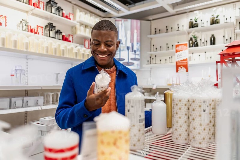 A young African man smiles while shopping in a store filled with various decorative candles. The shelves are stocked with colorful products