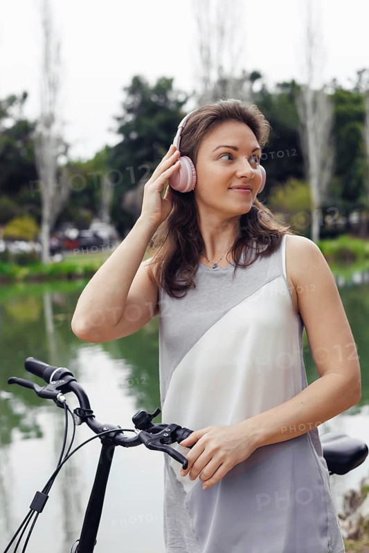 A young Caucasian woman with brown hair stands beside a bicycle near a serene lake. She wears pink headphones and smiles, enjoying the outdoors