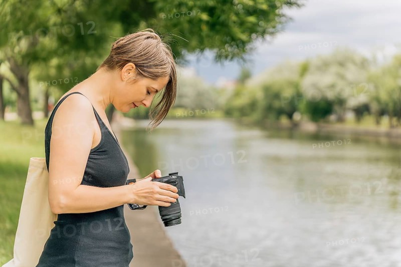 Woman 40s reviewing travel photos on camera by urban river, Woman examining her camera by a lakeside path lined with trees and greenery