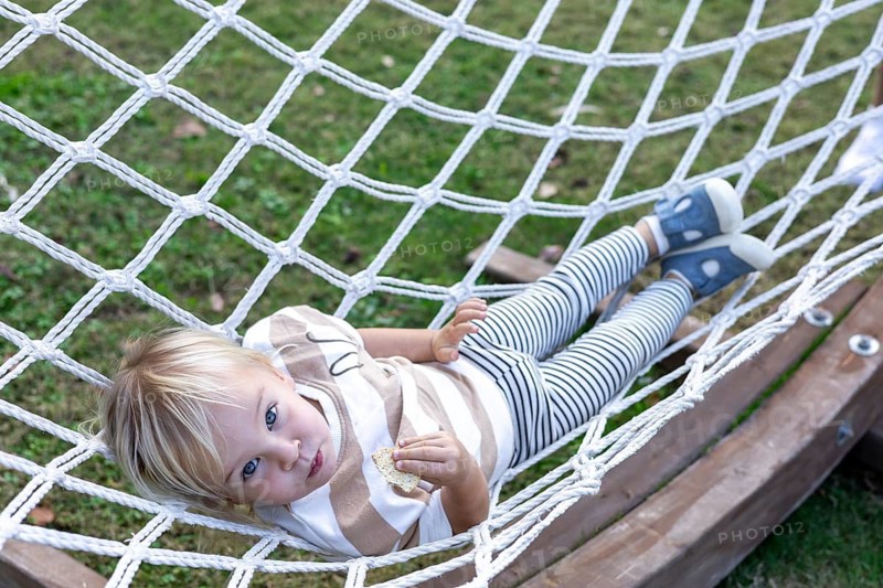 A young child enjoys a moment of relaxation in a hammock in backyard. Surrounded by nature, the scene showcases a tranquil environment ideal for leisure. Smiling toddler blondie girl