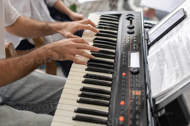 A close-up of hands playing a keyboard in a home setting. Sheet music is visible. The atmosphere is relaxed and intimate, suggesting a family concert