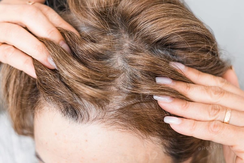 Mature middle-aged caucasian woman touching her gray, silver roots hair close-up on gray tiles background, hair loss and age-related changes, naturalness and natural aging concept