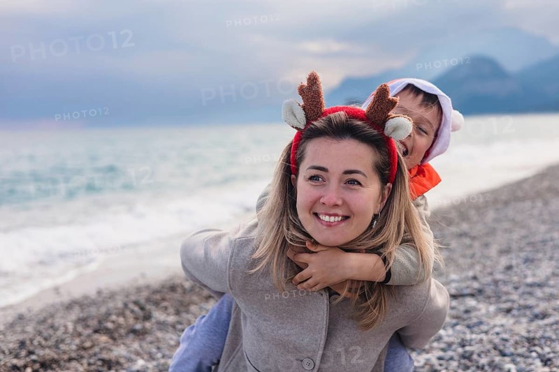A young Caucasian woman with long blonde hair wears reindeer antlers. A young boy with brown hair playfully rides on her back at a winter beach