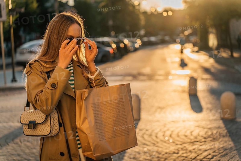 A woman walks around the city after shopping at sunset with a paper bag, Woman wearing sunglasses and a trench coat carries a bag on a sunlit street at sunset, Vilnius, Lithuania