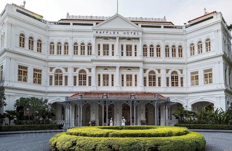 White colonial facade of the 1887 Raffles Hotel beside its circular lawn with staff at the entrance, Singapore