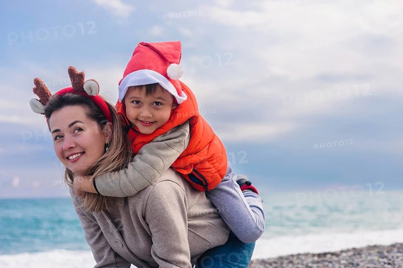 A young Caucasian woman with brown hair wears reindeer antlers. A young Asian boy in a Santa hat smiles while riding on her back at the beach during winter