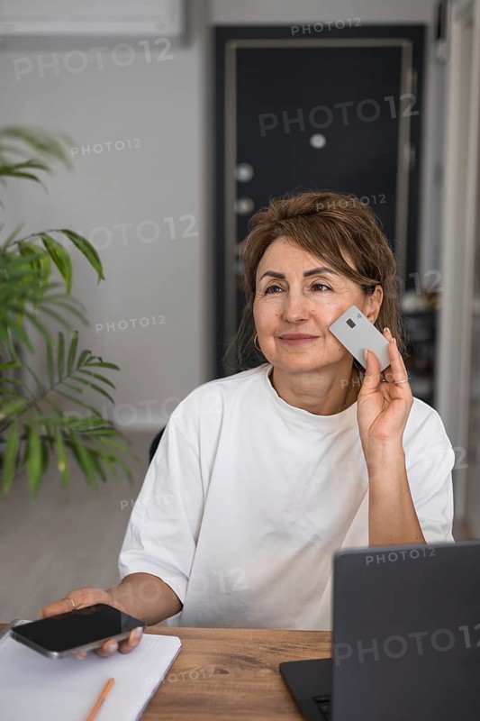 Pretty mature caucasian woman buying goods and services, making order on-line using laptop, smartphone and credit card at home, e-mailing, solve business remotely. Online mobile application shopping