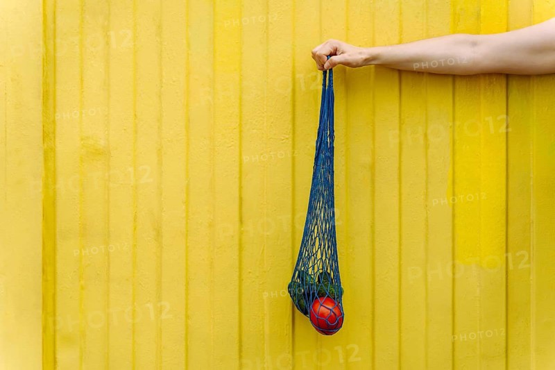 Eco-friendly mesh tote with fresh veggies isolated on yellow background, A hand holding a blue net bag with a red ball against a yellow wall