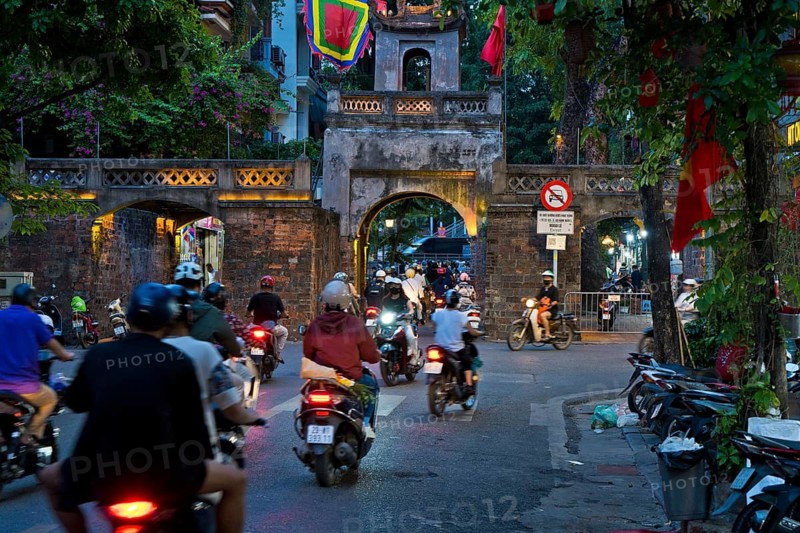 City gate during the evening rush hour on a busy street with mopeds, scooters, motorbikes and passers-by, surrounded by historic buildings and trees, Historic City Gate in the Old Quarter of Hanoi Vietnam