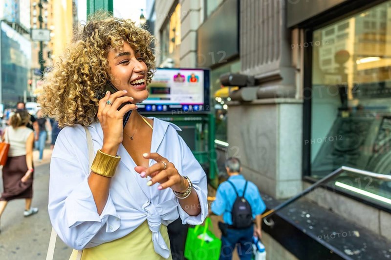 Young curly haired woman enjoying a phone call and laughing, walking on a busy manhattan street, embracing urban lifestyle and technology