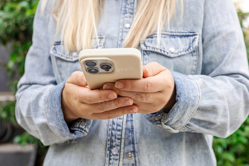 A young Caucasian woman with long blonde hair holds a smartphone in her hands. She wears a denim shirt and stands in a green outdoor setting