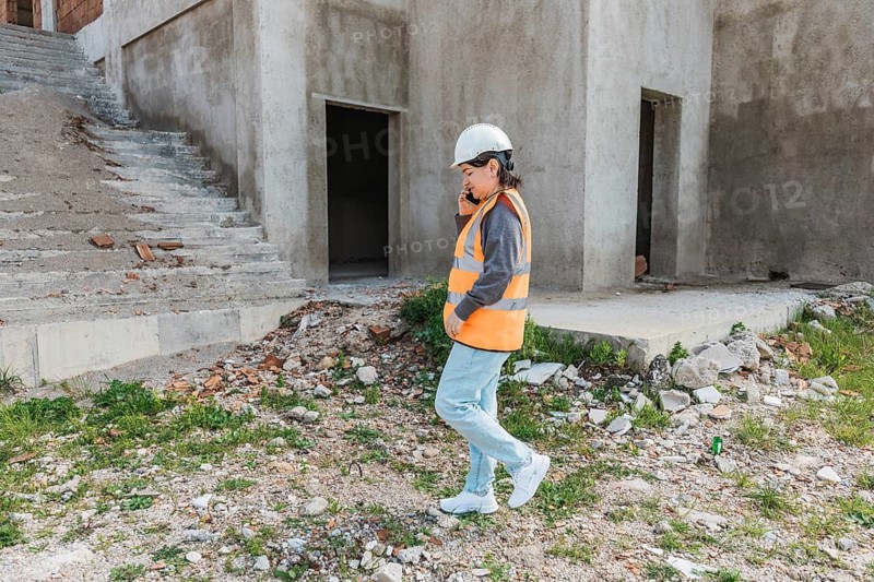 A young Hispanic woman in a safety vest and hard hat walks on a construction site. The background features unfinished concrete structures and debris