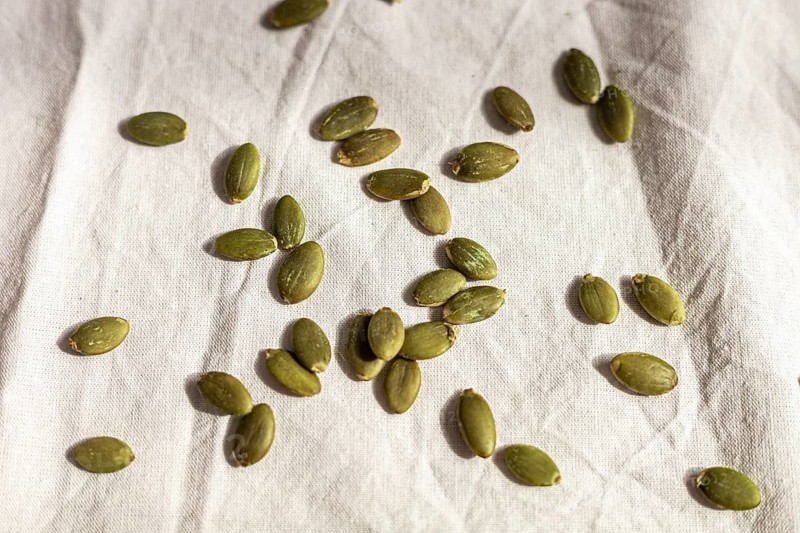 Green pumpkin seeds scattered on a light fabric background. The seeds are raw and unroasted, showcasing their natural color and texture