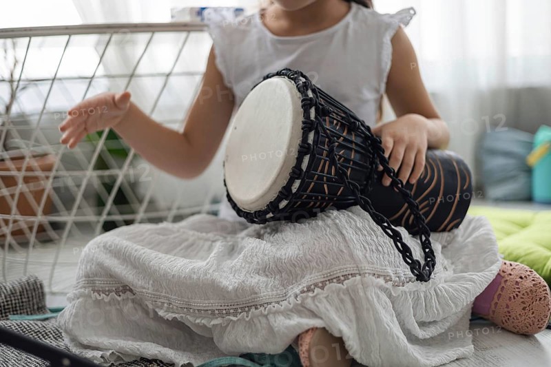A young girl plays a black drum at home. She wears a white dress and sits on a colorful mat. The room is bright and cozy, filled with family warmth