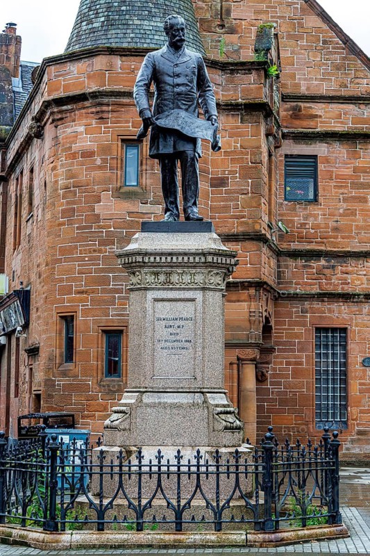 Blackened bronze statue of shipbuilder Sir William Pearce holding a design plan, standing at Govan Cross opposite the Pearce Institute, Glasgow, Scotland, United Kingdom