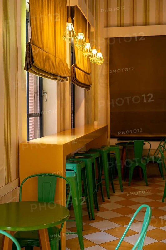 This cozy cafe features a bright yellow countertop with green seating. Warm lighting hangs from the ceiling, creating an inviting atmosphere. Large windows let in natural light