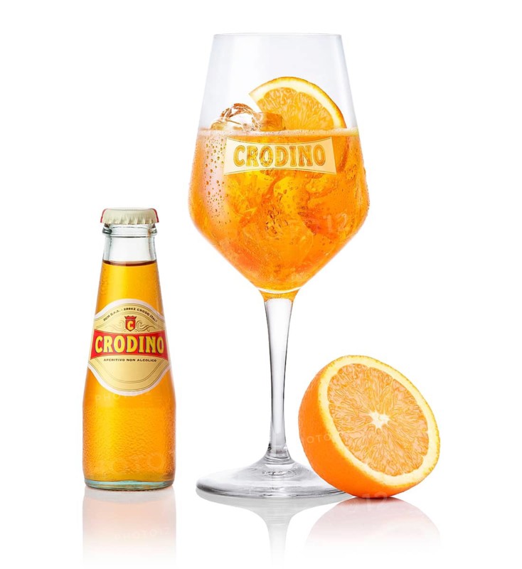 Crodino on ice cube cubes in a branded wine glass, half an orange slice and half orange as a garnish, next to a bottle of crodino, in front of a white background, studio shot