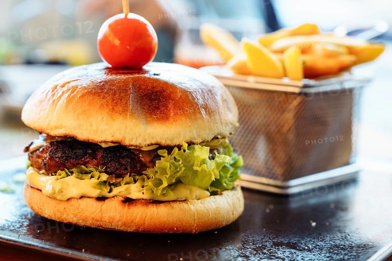 Focus on the foreground burger and fries, A close-up of a gourmet burger with lettuce and a cherry tomato on top, accompanied by fries in a metal basket