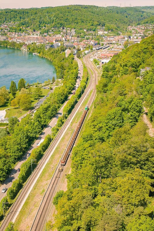 River bank with railway line and surrounding forest, city in the background, summery atmosphere, construction site by Leonhard Weiß to renovate the train station and build the overhead line, Waldshut, Germany