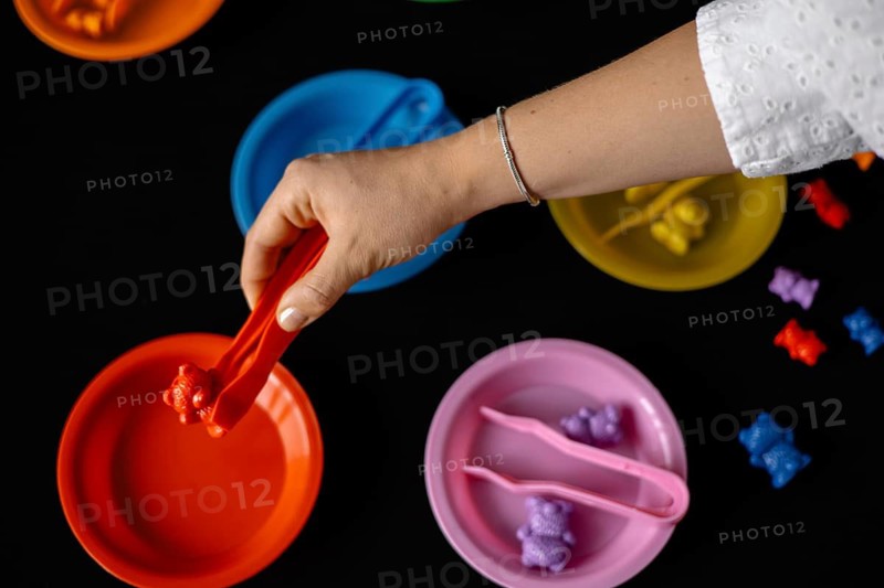 A hand sorts bears using tongs into colorful plates on a dark surface