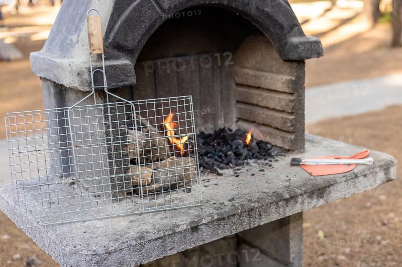 A stone barbecue grill with burning charcoal and flames. A metal grill rack is placed on top. The setting is outdoors with a natural background