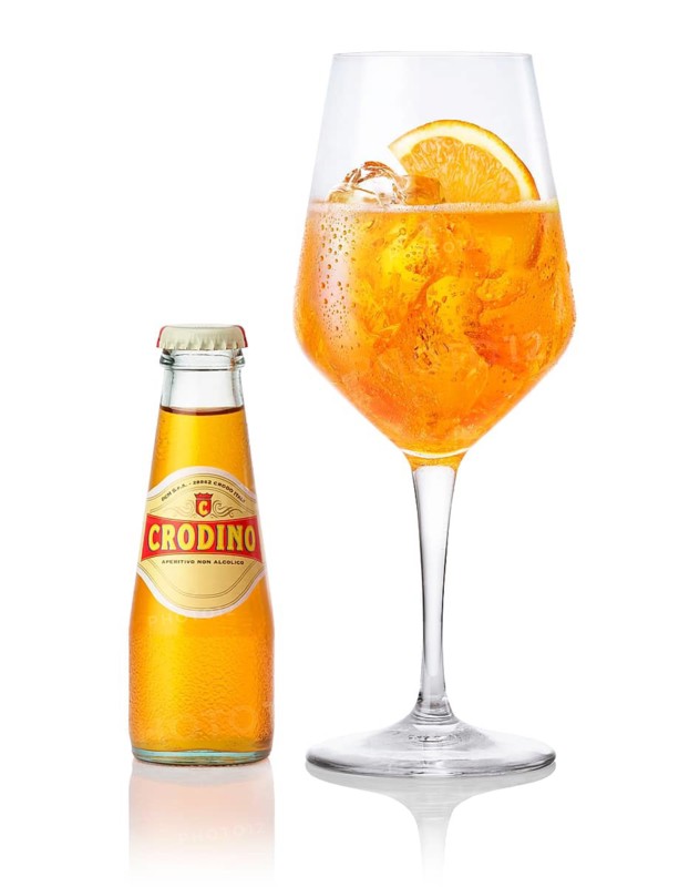 Crodino on ice cube cubes in a wine glass, half an orange slice as garnish, next to a bottle of crodino, in front of a white background, studio shot
