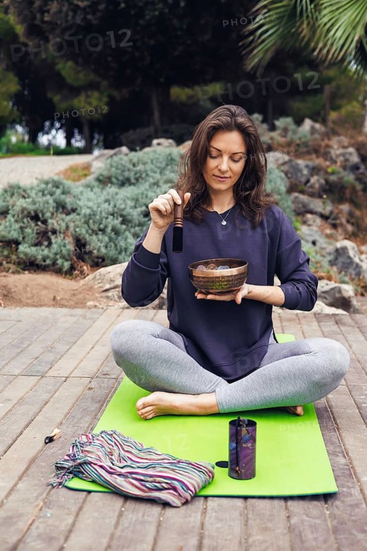 A young Caucasian woman with brown hair plays a singing bowl outdoors. She wears a dark sweater and sits on a mat in a serene garden setting