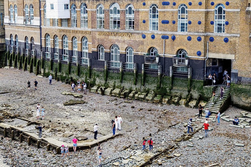 At low tide, families and mudlarkers pick across the stone and shingle foreshore below Horselydown Old Stairs, a historic watermen's landing recorded on eighteenth century maps, with the lettered facade of the former Anchor Brewhouse rising behind, London, England, United Kingdom