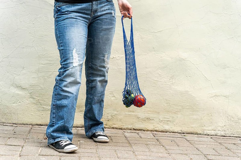 Urban grocery run: blue mesh bag with veggies in denim and sneakers POV, Person holding a blue mesh bag with items in front of a textured concrete wall